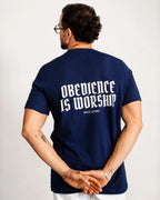 A man viewed from behind wearing a navy blue t-shirt with 'OBEDIENCE IS WORSHIP' printed in white gothic-style lettering on the back, with 'HOLY LOOKS' underneath. He is standing against a plain light gray background.