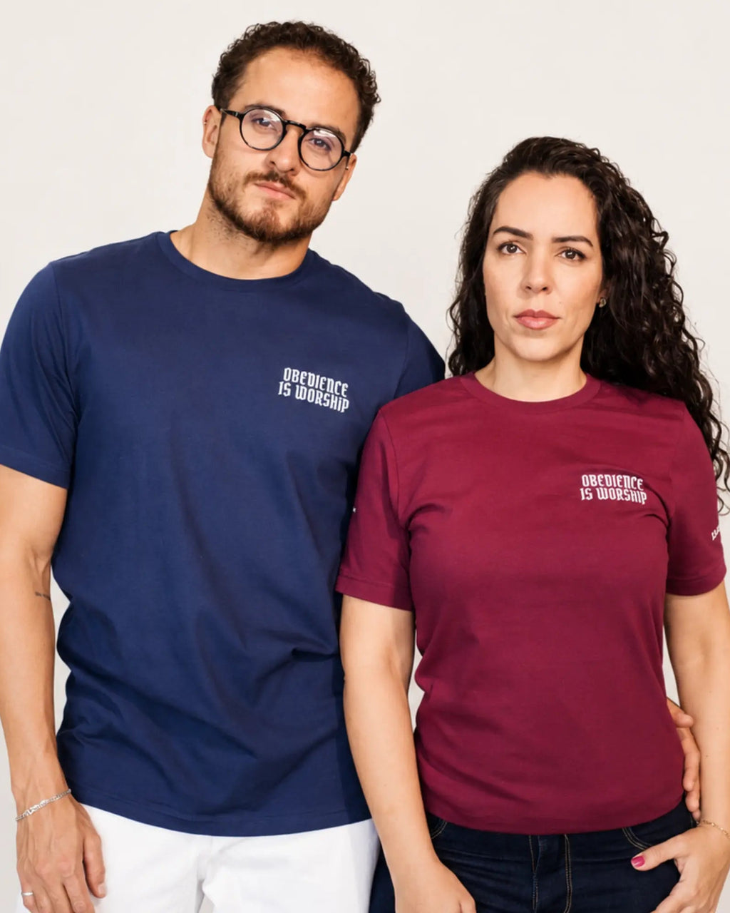 A man and a woman standing side-by-side wearing matching graphic t-shirts with the text 'OURDEST IS OWEASP' printed in white on the chest. The man wears a navy blue t-shirt and white pants, while the woman wears a burgundy t-shirt and dark jeans, set against a plain light gray background.