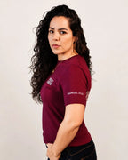 A woman with long, curly dark hair is shown from the waist up, wearing a short-sleeve maroon t-shirt with white text on the sleeve and chest, paired with dark jeans, against a plain light background.