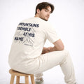 Man wearing a white long-sleeve shirt with text and graphics on the back, sitting on a wooden stool against a white background.