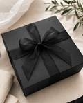 A square black gift box wrapped with a wide black ribbon and bow, styled on a light beige fabric surface with soft white tissue paper and green leaves in the background.
