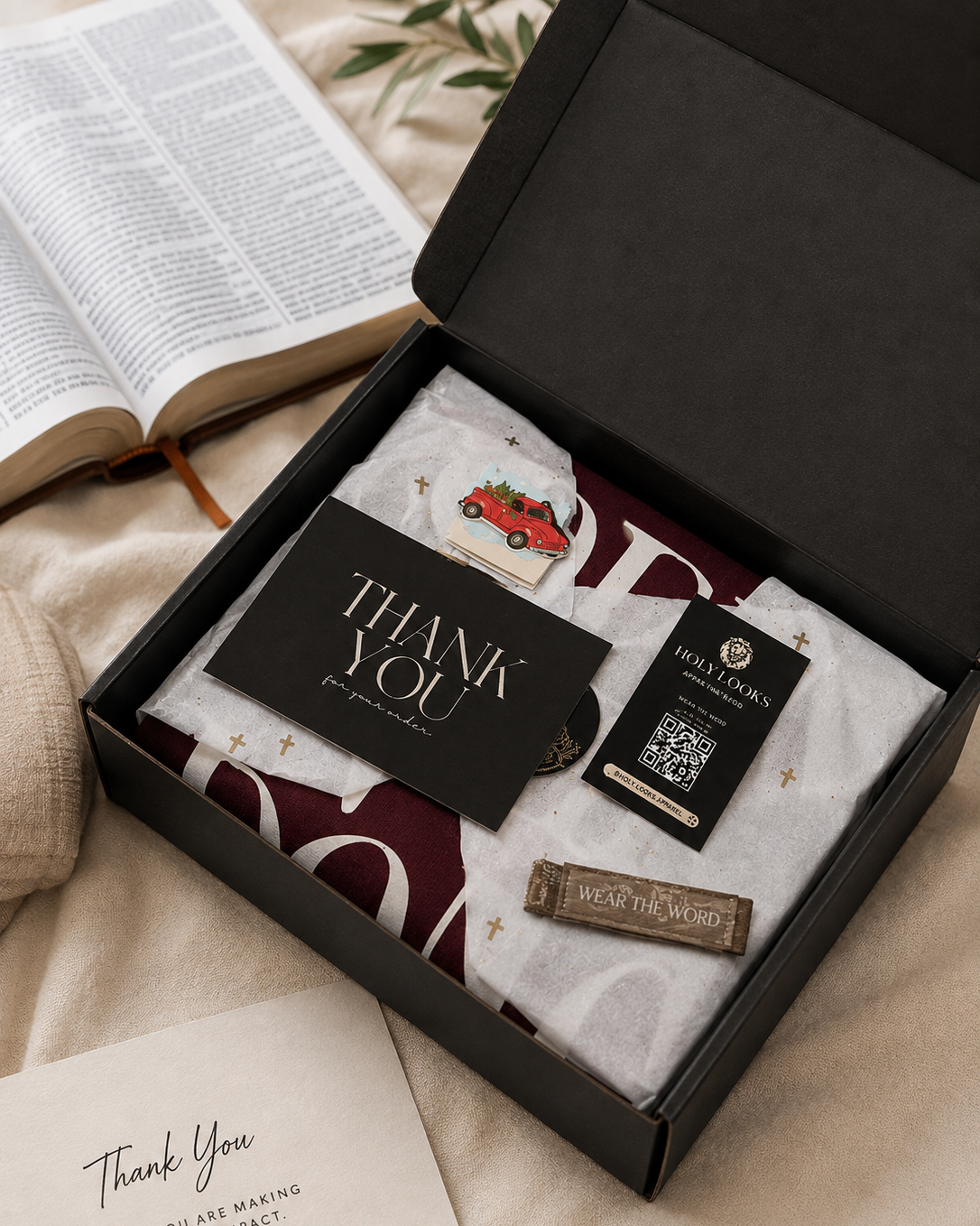 A black gift box opened on a beige fabric surface, containing folded apparel with a burgundy and white pattern, a black thank-you card, a small card reading 'WE LOVE YOU', and a wooden tag. An open book and a second thank-you card are visible around it.