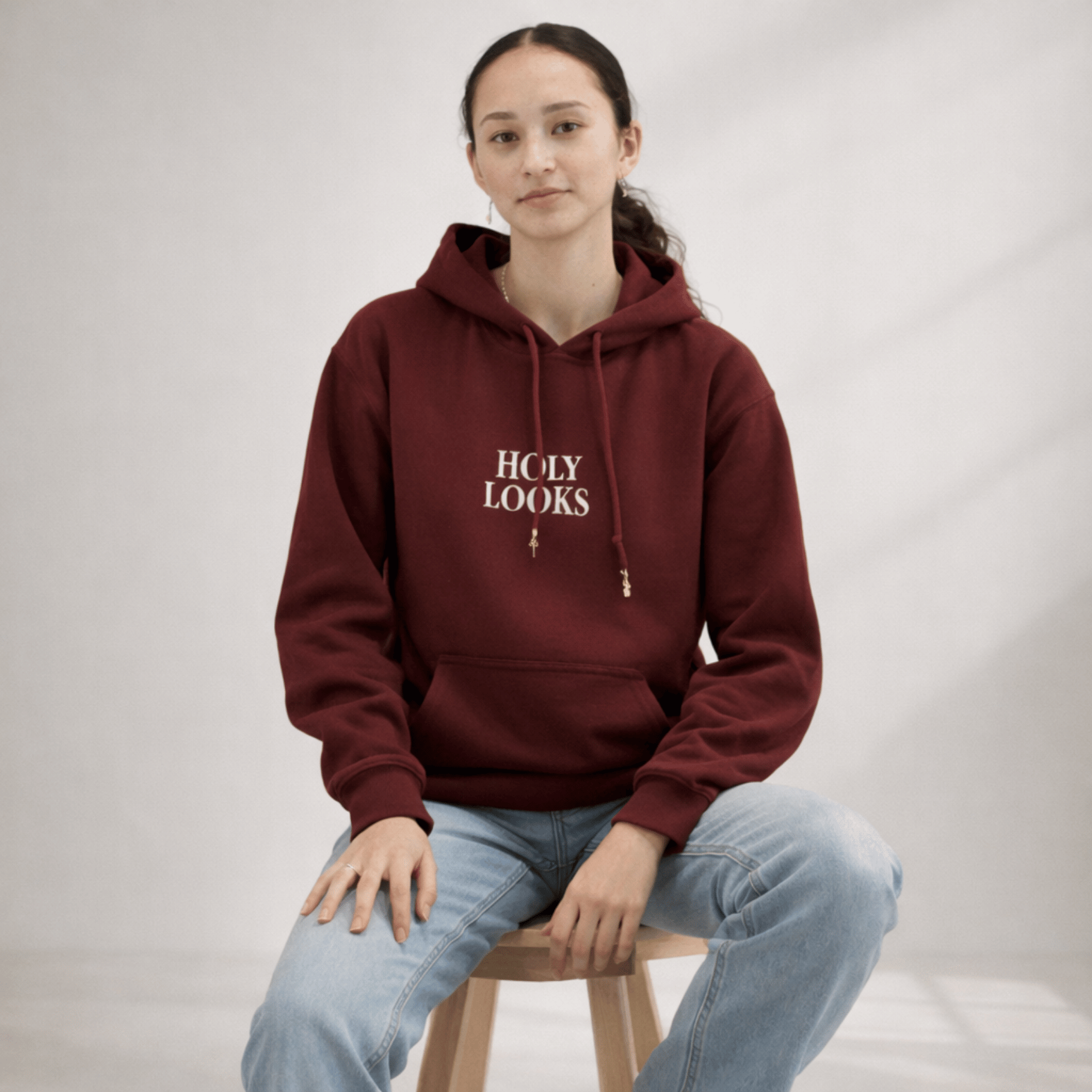 Person wearing a maroon hoodie with 'HOLY LOOKS' text, sitting on a stool against a plain background