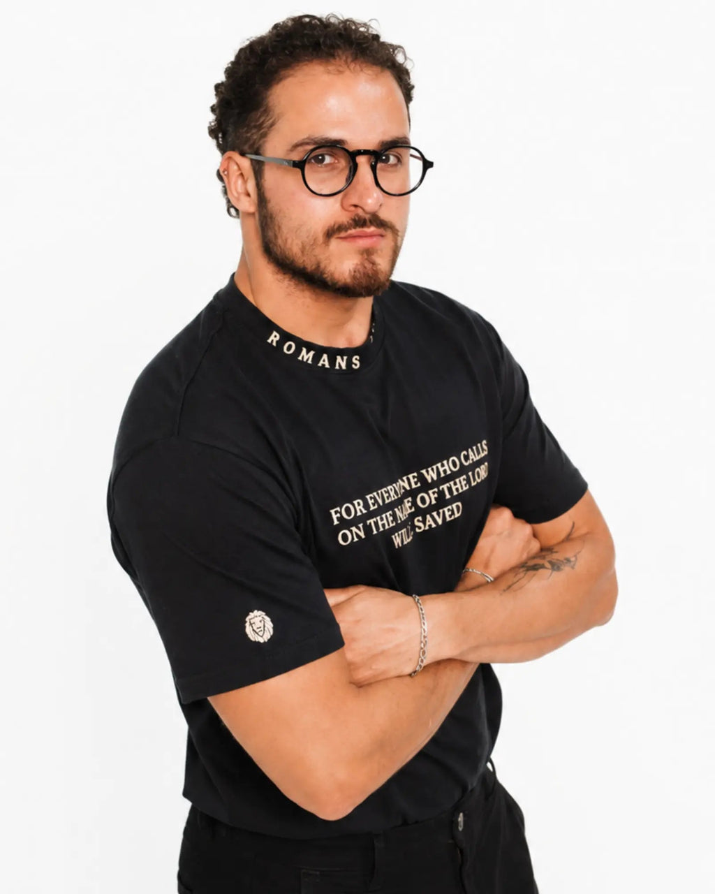 A man wearing a black graphic t-shirt with white text, standing against a plain white background with his arms crossed.