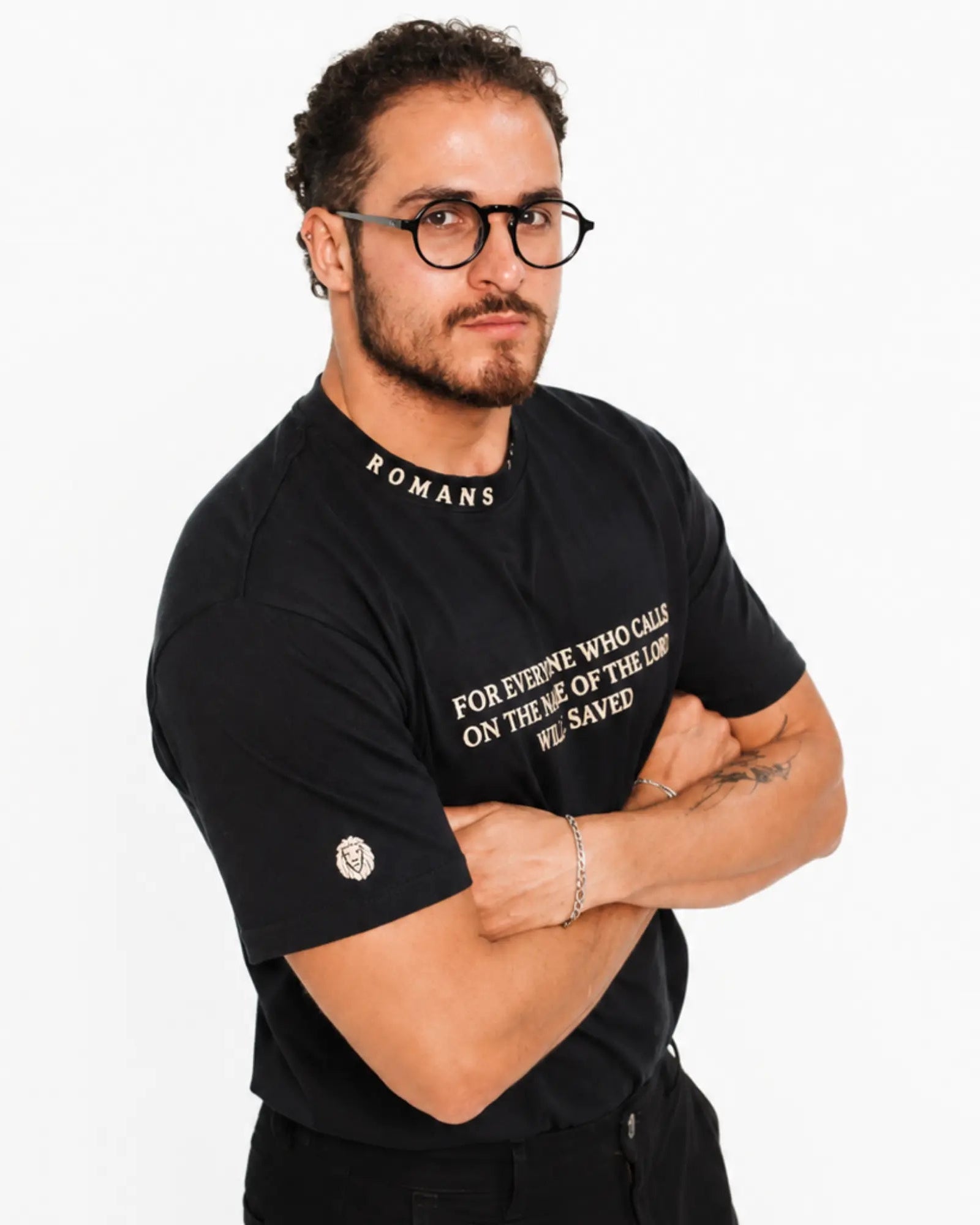 A man wearing a black graphic t-shirt with white text, standing against a plain white background with his arms crossed.