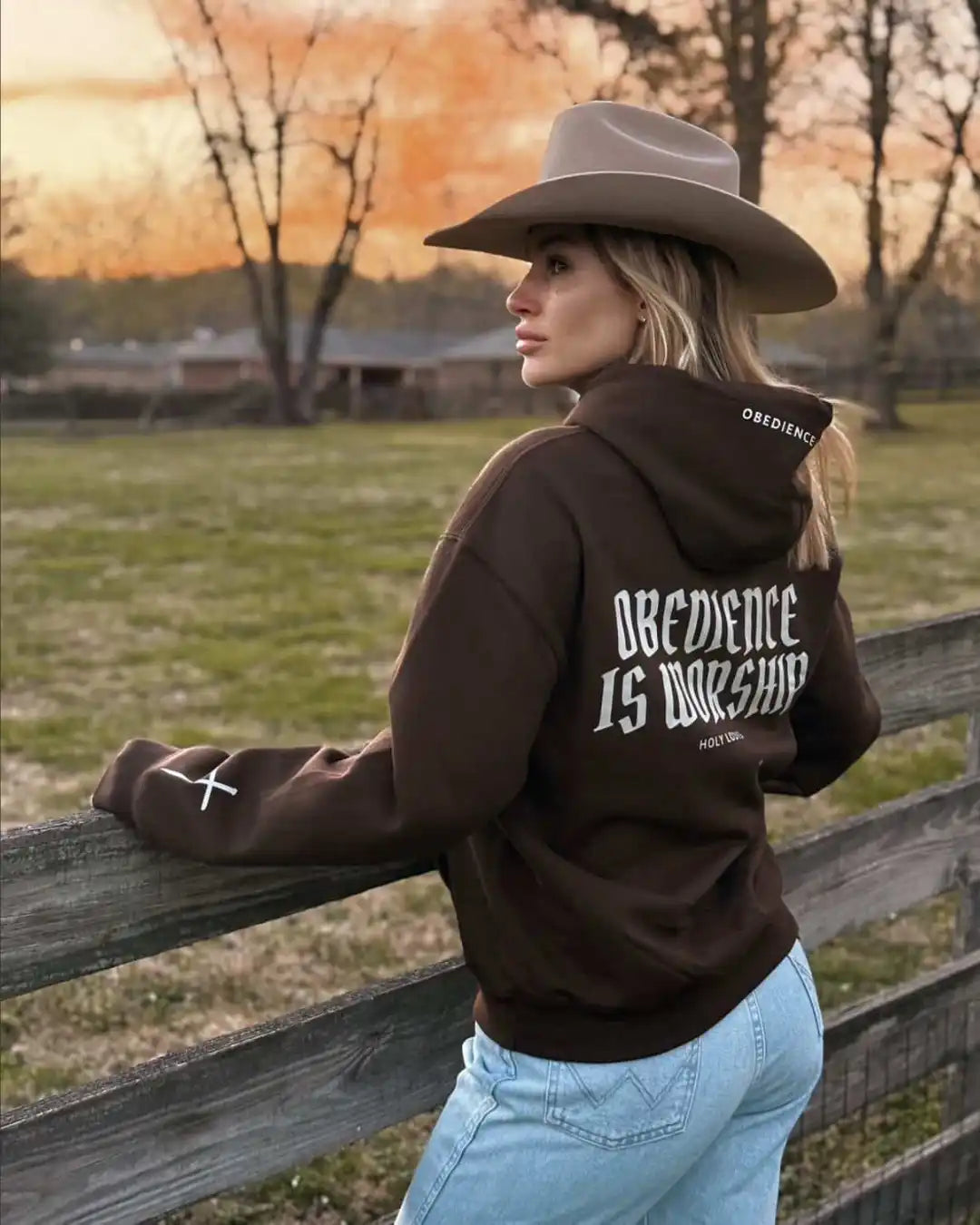 Person wearing a hoodie with 'Obedience is Worship' text, standing by a wooden fence with a sunset in the background.