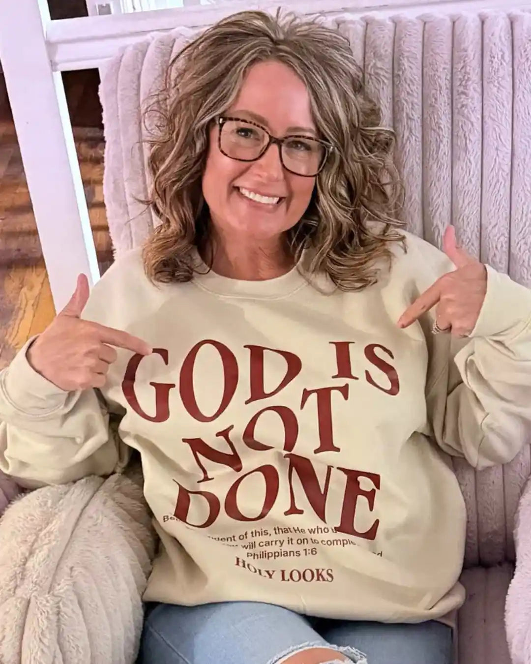 Woman wearing a beige sweatshirt with 'God is not done' text, sitting on a couch.