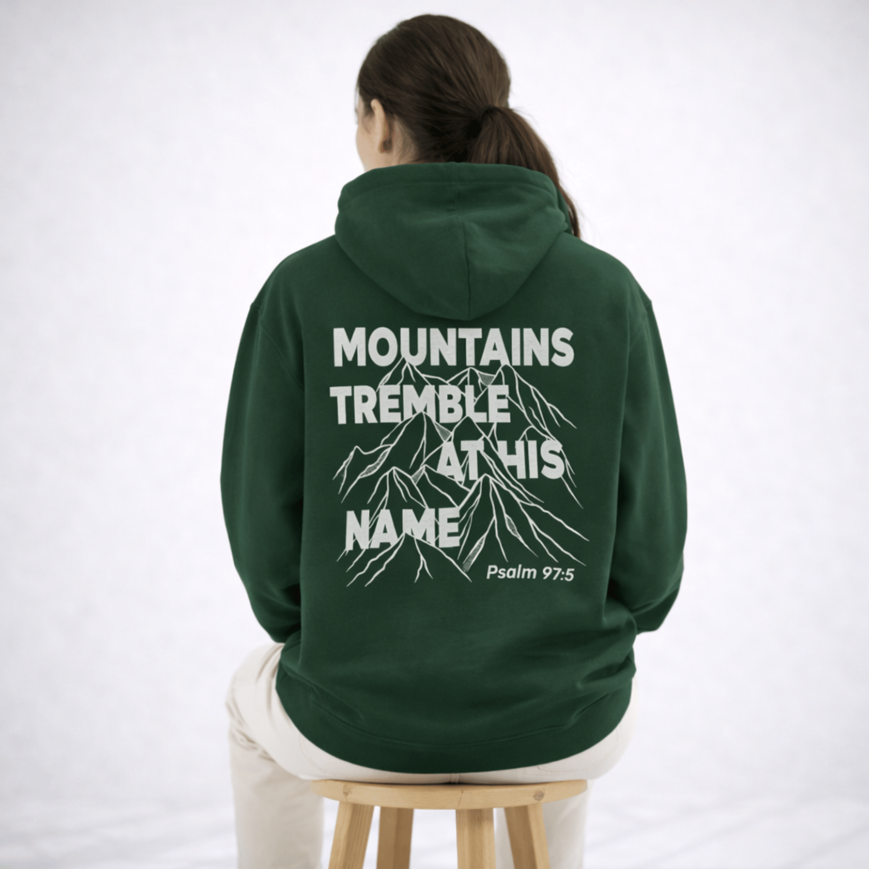 Person wearing a green hoodie with text and mountain graphic on the back, sitting on a stool against a white background.