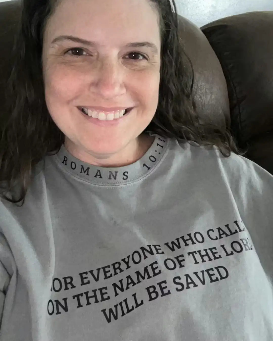 Person wearing a gray t-shirt with a religious quote, sitting on a brown couch.