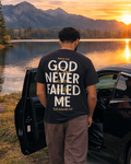 A man stands with his back to the camera, next to an open black car door, wearing a black t-shirt with the phrase 'GOD NEVER FAILED ME' printed in white on the back. He is set against a scenic background of a mountain lake at sunset.