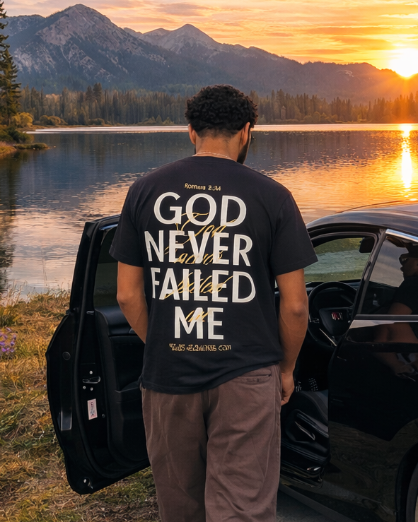 A man stands with his back to the camera, next to an open black car door, wearing a black t-shirt with the phrase 'GOD NEVER FAILED ME' printed in white on the back. He is set against a scenic background of a mountain lake at sunset.
