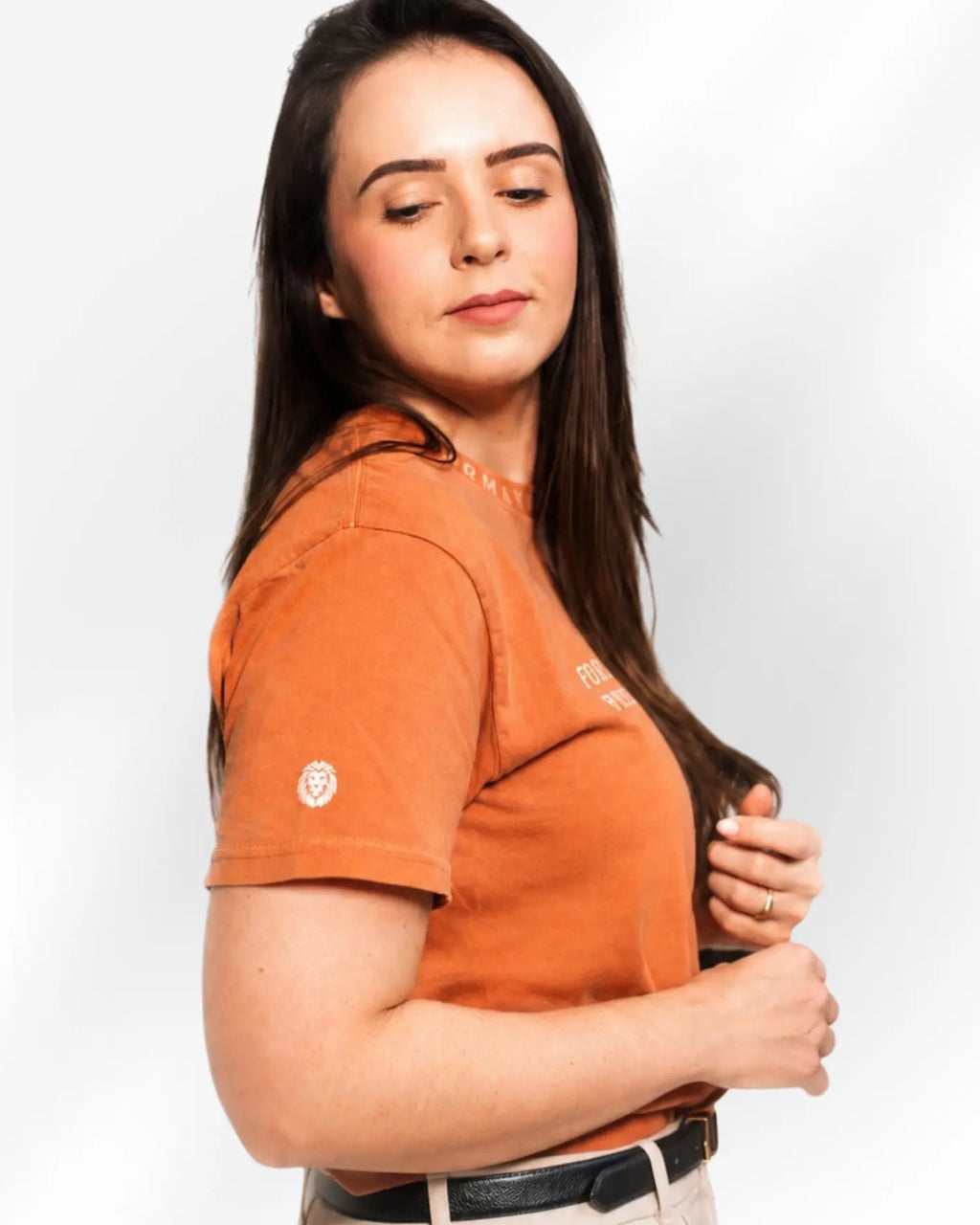 A woman wearing a short-sleeve orange t-shirt and beige trousers, shown from the waist up against a light gray studio background.