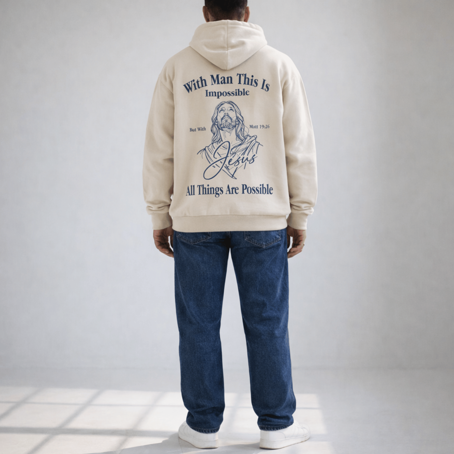 Person wearing a beige hoodie with text and a graphic of a religious figure on the back, standing against a plain background.