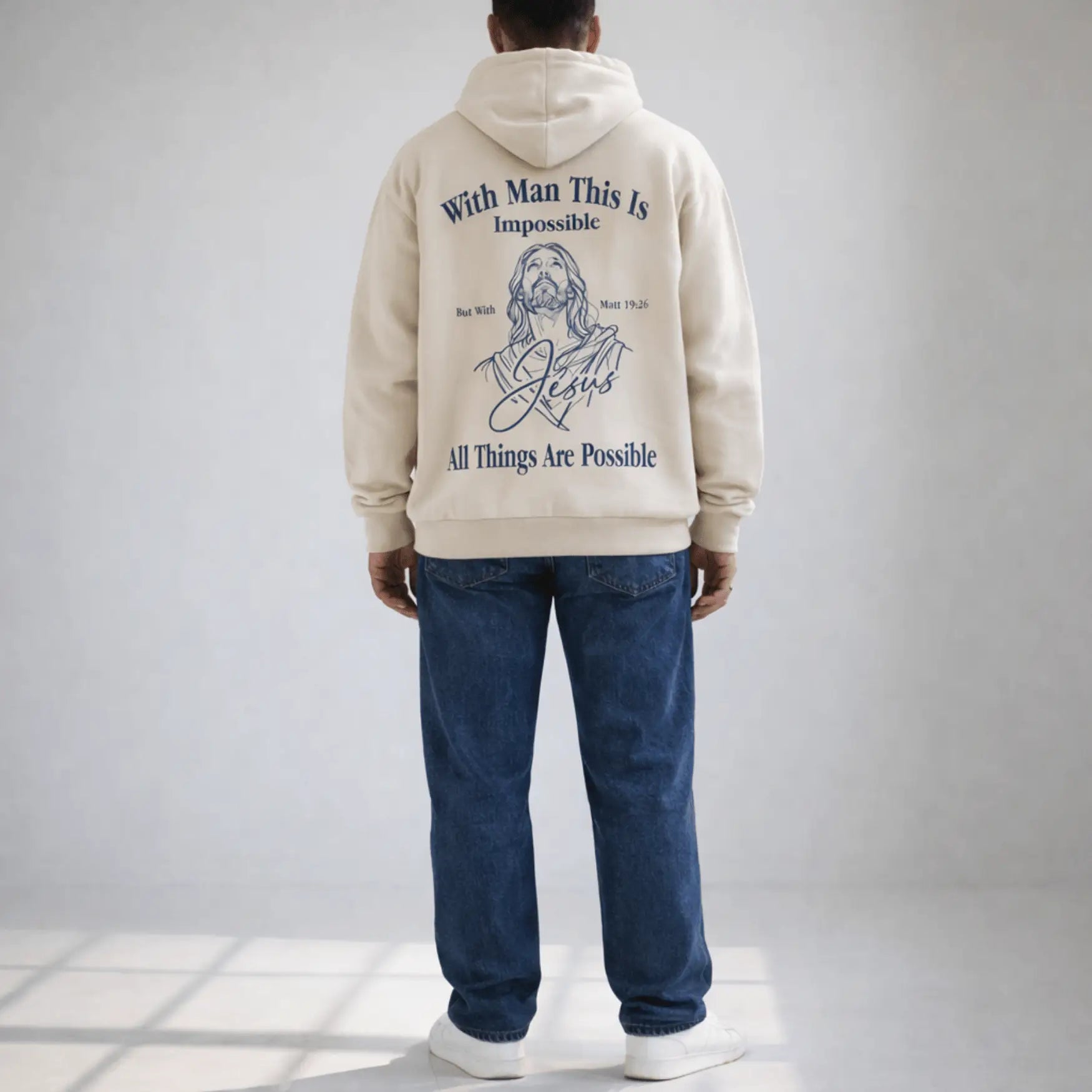 Person wearing a beige hoodie with text and a graphic of a religious figure on the back, standing against a plain background.