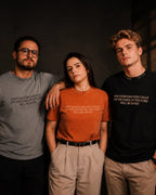Three people standing together in a dim indoor setting, each wearing a graphic T-shirt with white printed text. The man on the left wears a gray T-shirt, the woman in the center wears an orange T-shirt, and the man on the right wears a black T-shirt.