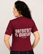 A woman shown from the back wearing a maroon short-sleeve t-shirt with 'OBEDIENCE IS WORSHIP' printed in white gothic-style lettering on the back, paired with dark jeans against a light gray studio background.