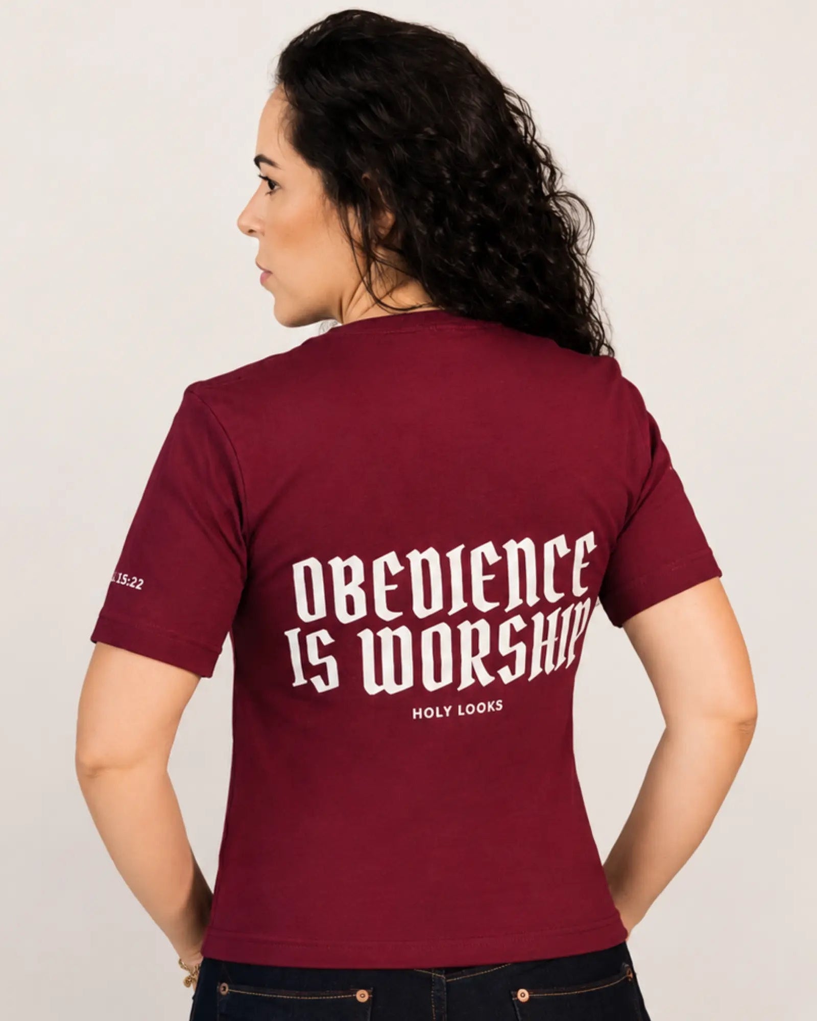 A woman shown from the back wearing a maroon short-sleeve t-shirt with 'OBEDIENCE IS WORSHIP' printed in white gothic-style lettering on the back, paired with dark jeans against a light gray studio background.