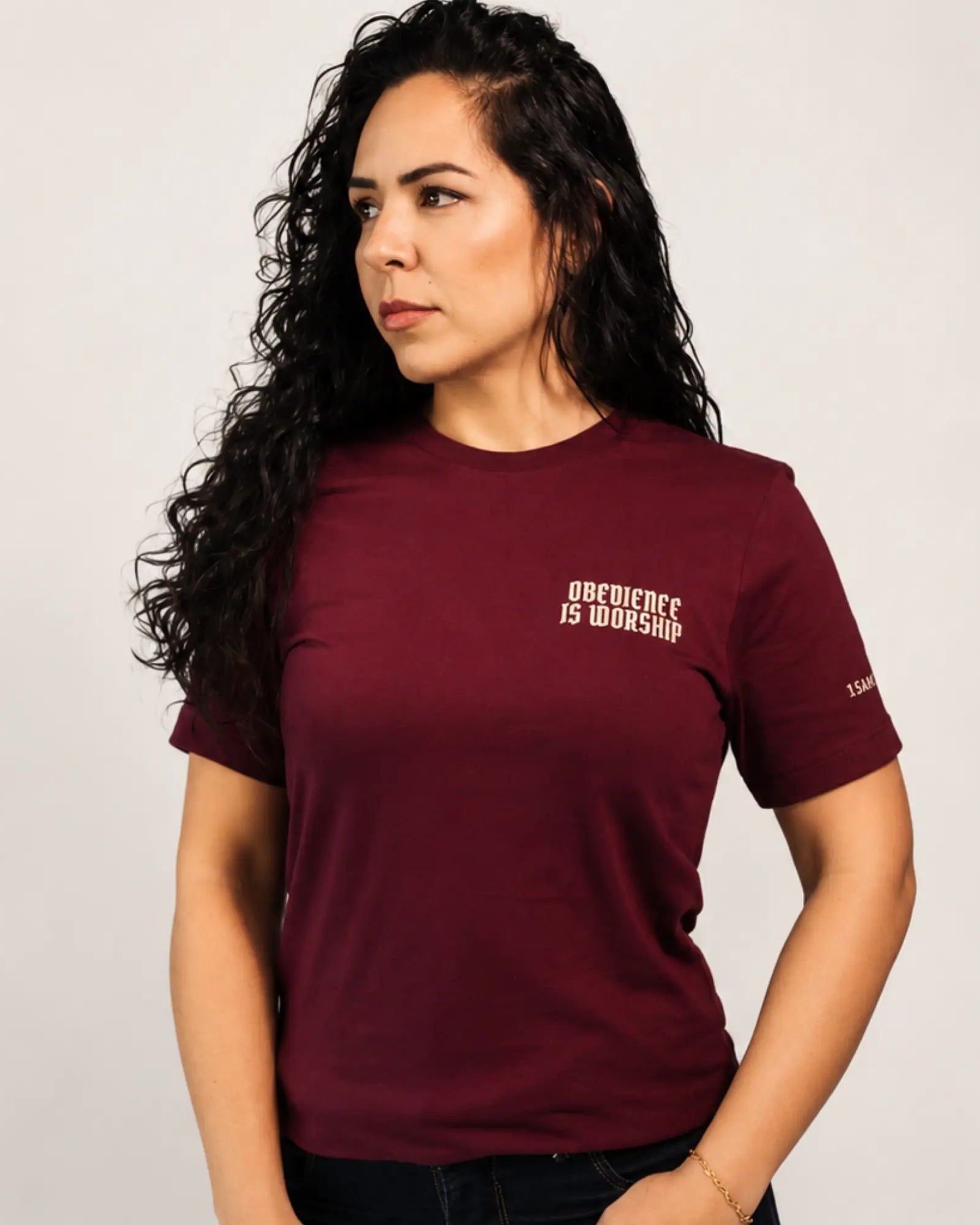 A woman with long, curly dark hair wearing a maroon short-sleeve t-shirt with white text on the left chest that reads 'OBEYIENCE IS WORSHIP' and a small white logo on the right sleeve. She is posing against a light gray studio background.