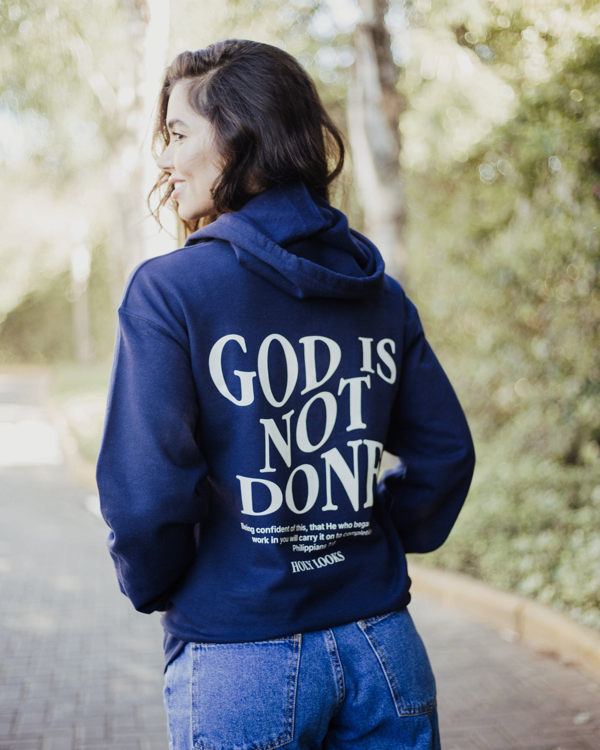 A woman wearing a navy blue hoodie with 'GOD IS NOT DONE' printed in white on the back, standing on an outdoor paved path with trees in the background.