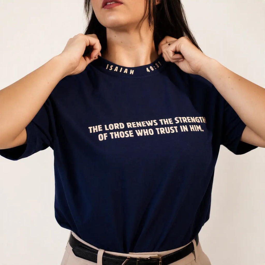 A woman wearing a navy blue t-shirt with white printed text from Isaiah 46:3 on the chest, holding the collar of the shirt. The background is a clean, off-white studio wall.