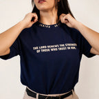 A woman wearing a navy blue t-shirt with white printed text from Isaiah 46:3 on the chest, holding the collar of the shirt. The background is a clean, off-white studio wall.