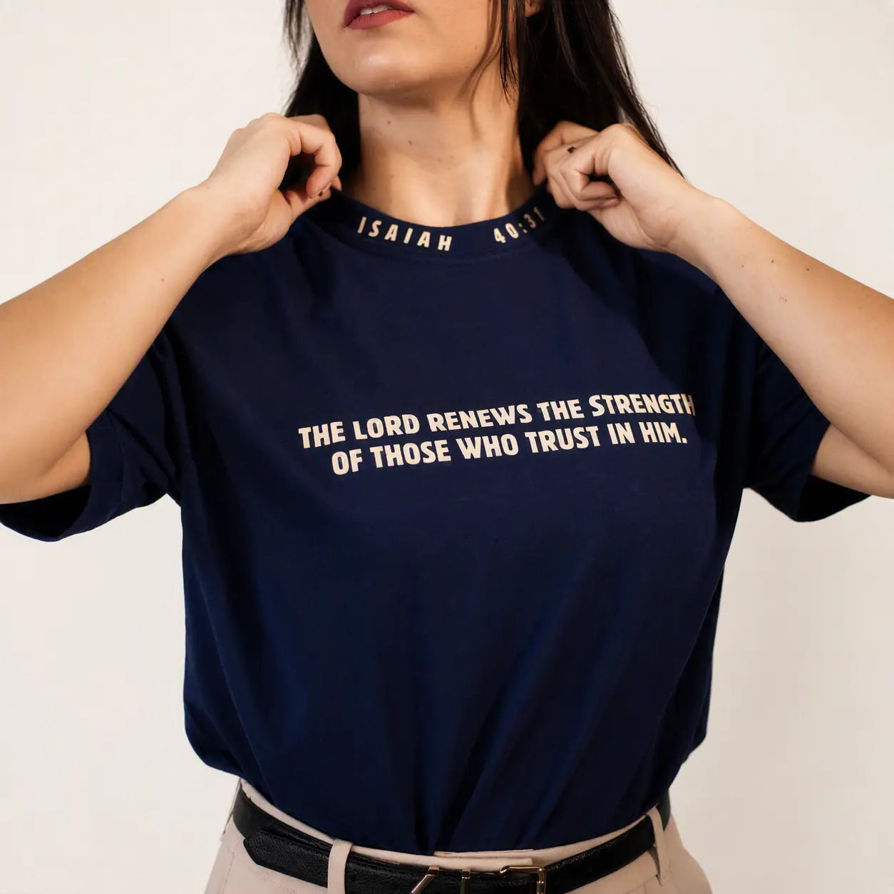 A woman wearing a navy blue t-shirt with white printed text from Isaiah 46:3 on the chest, holding the collar of the shirt. The background is a clean, off-white studio wall.