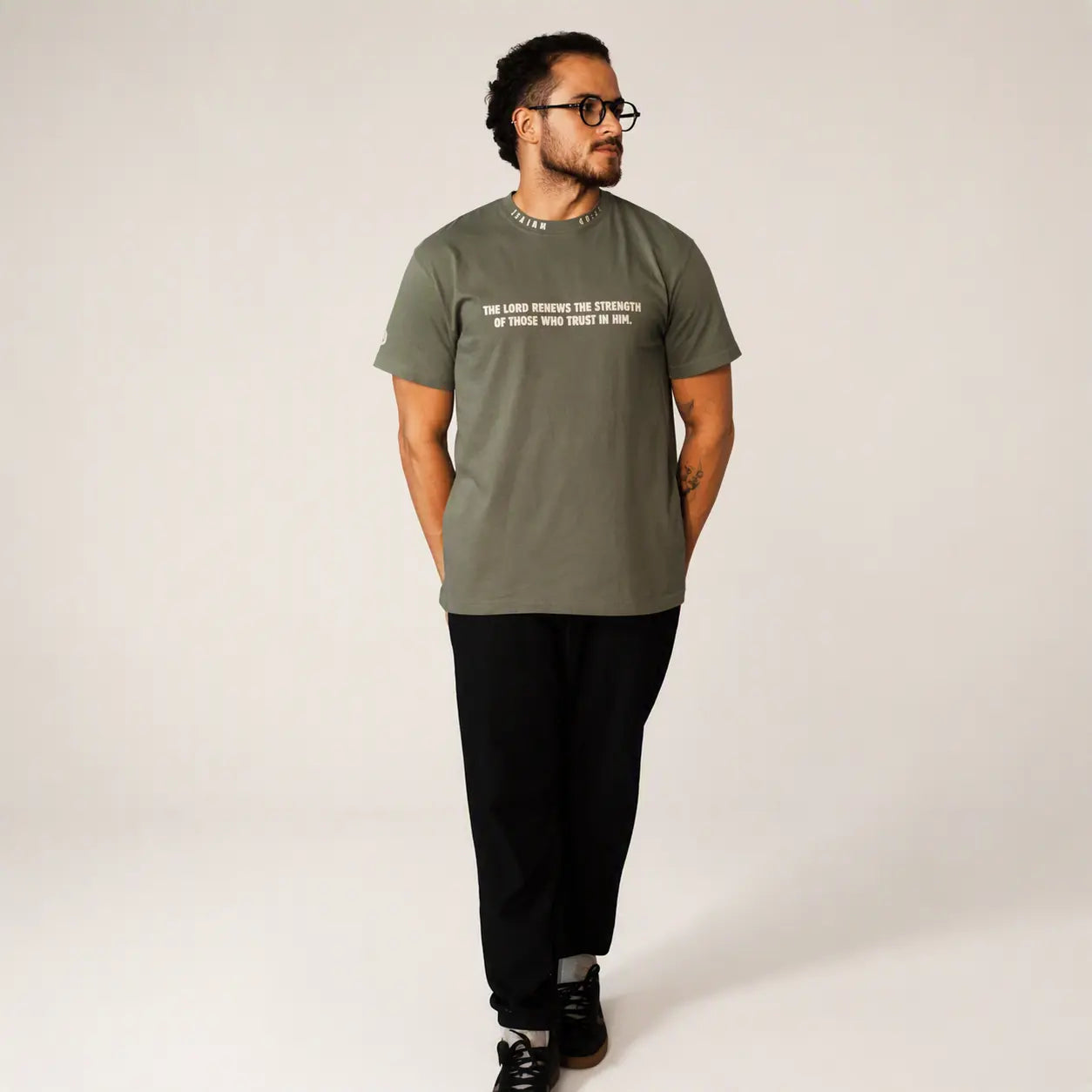 A man wearing a muted olive green graphic t-shirt with white text, paired with black pants and black sneakers, standing against a plain light gray studio background.