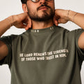 A man wearing an olive green crewneck t-shirt with a white printed bible verse on the chest, posing with his hands behind his neck.