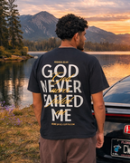 A man stands with his back to the camera, wearing a black graphic t-shirt that reads 'GOD NEVER FAILED ME' in white text, paired with brown pants. He is positioned next to a black Honda Civic in front of a scenic mountain lake sunset.