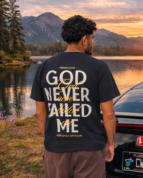 A man stands with his back to the camera, wearing a black graphic t-shirt that reads 'GOD NEVER FAILED ME' in white text, paired with brown pants. He is positioned next to a black Honda Civic in front of a scenic mountain lake sunset.
