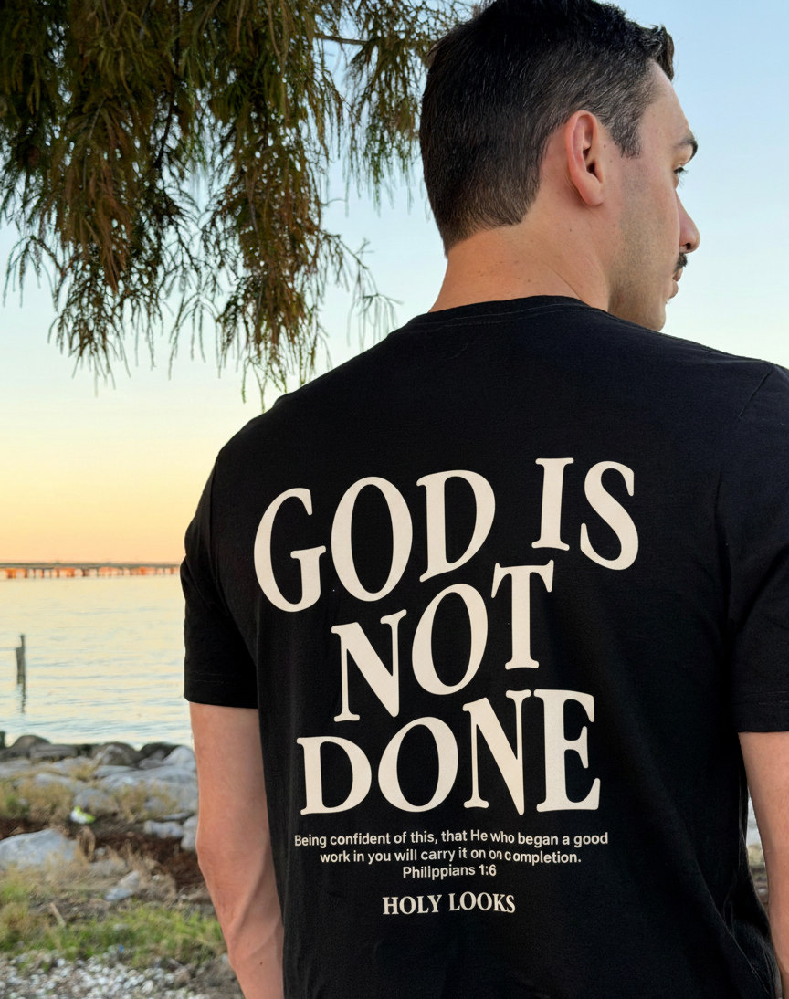 Man wearing a black t-shirt with 'GOD IS NOT DONE' text, standing outdoors.