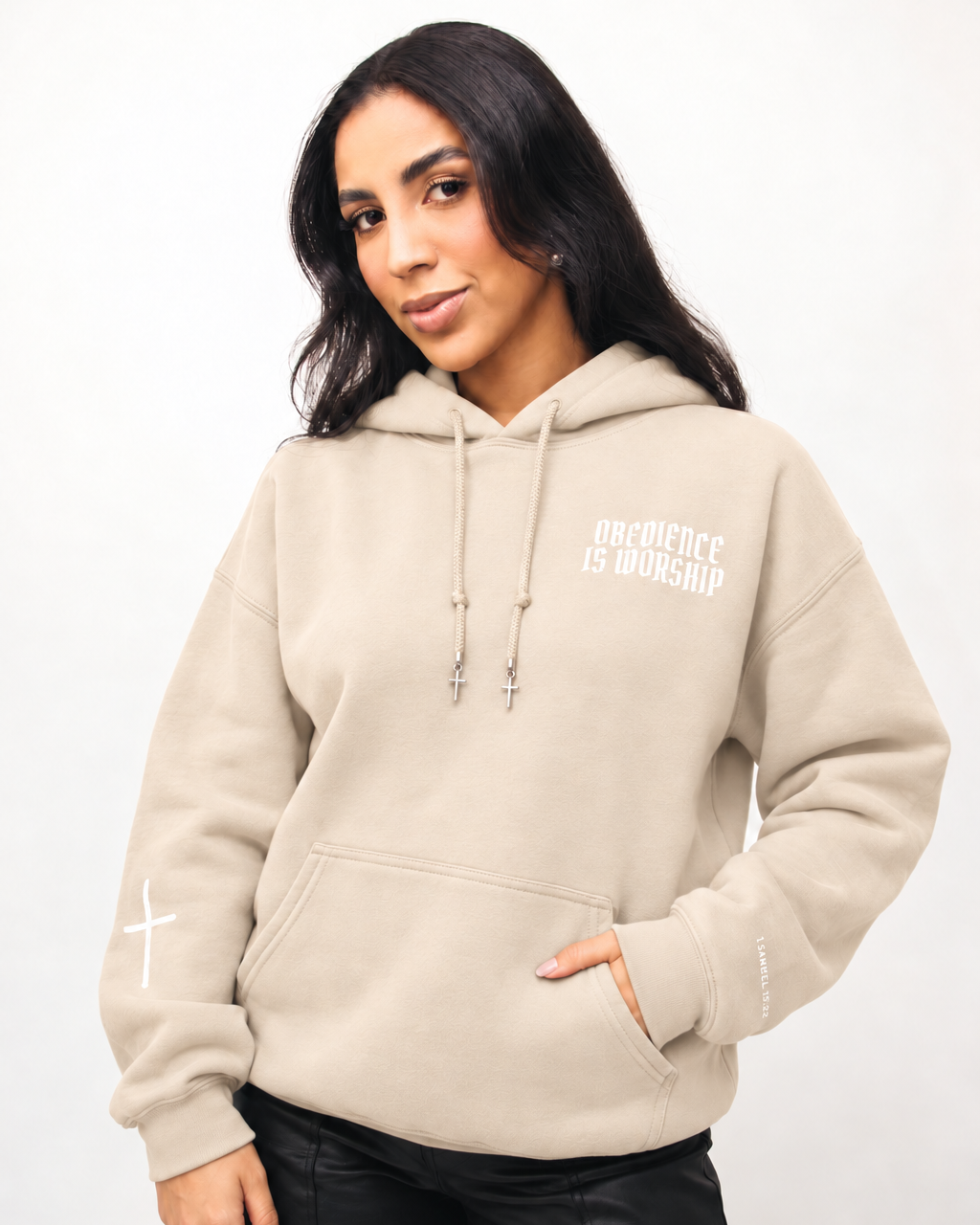 SAND A woman wearing a beige pullover hoodie with white graphic text reading 'ONE OFFENSE IS WORSHIP' on the left chest, paired with black pants, against a plain white studio background.