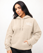 SAND A woman wearing a beige pullover hoodie with white graphic text reading 'ONE OFFENSE IS WORSHIP' on the left chest, paired with black pants, against a plain white studio background.