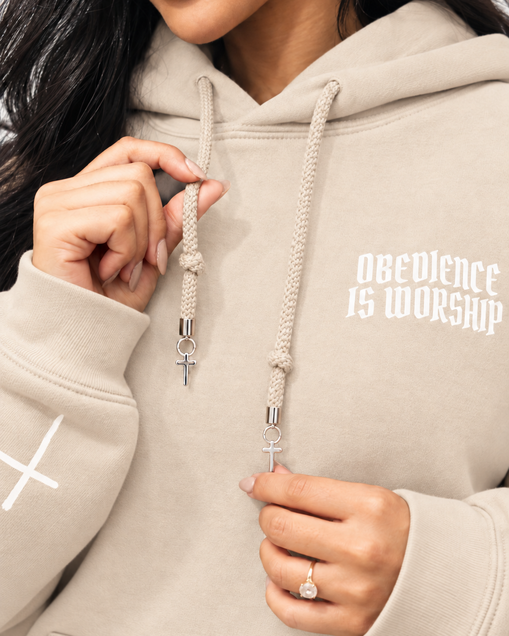 SAND Close-up of a person wearing a light beige hoodie with white text reading 'OBEYENCE IS WORSHIP' on the chest and white rope drawstrings with silver cross charms.