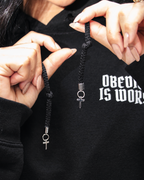 BLACK A close-up of a person holding a black braided cord necklace with silver-toned metal charms, including a ring and a cross, against a black hoodie with white text.