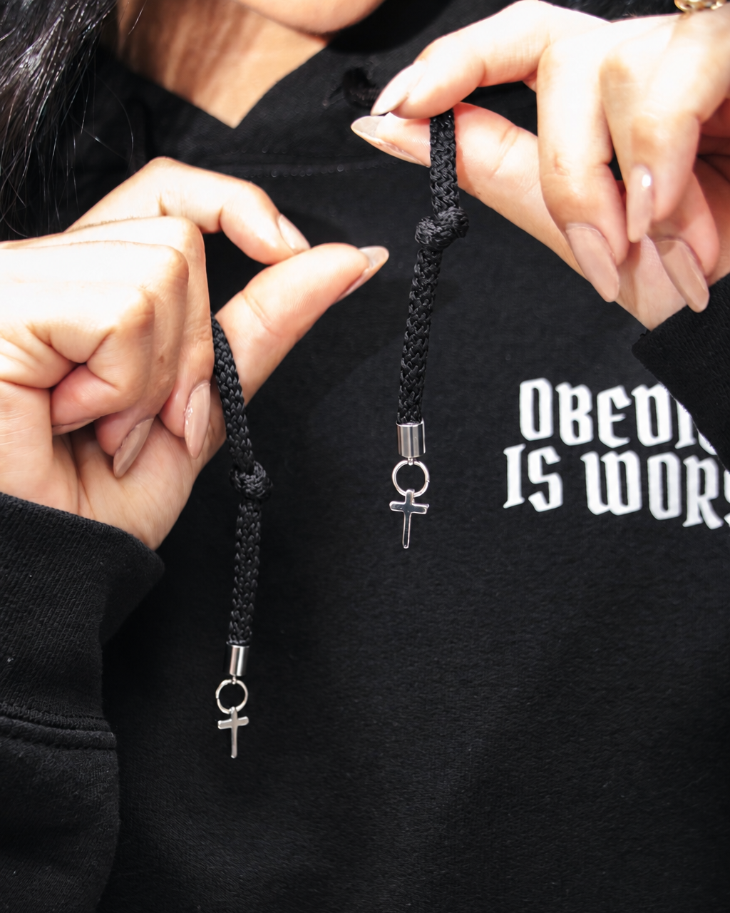 BLACK A close-up of a person holding a black braided cord necklace with silver-toned metal charms, including a ring and a cross, against a black hoodie with white text.