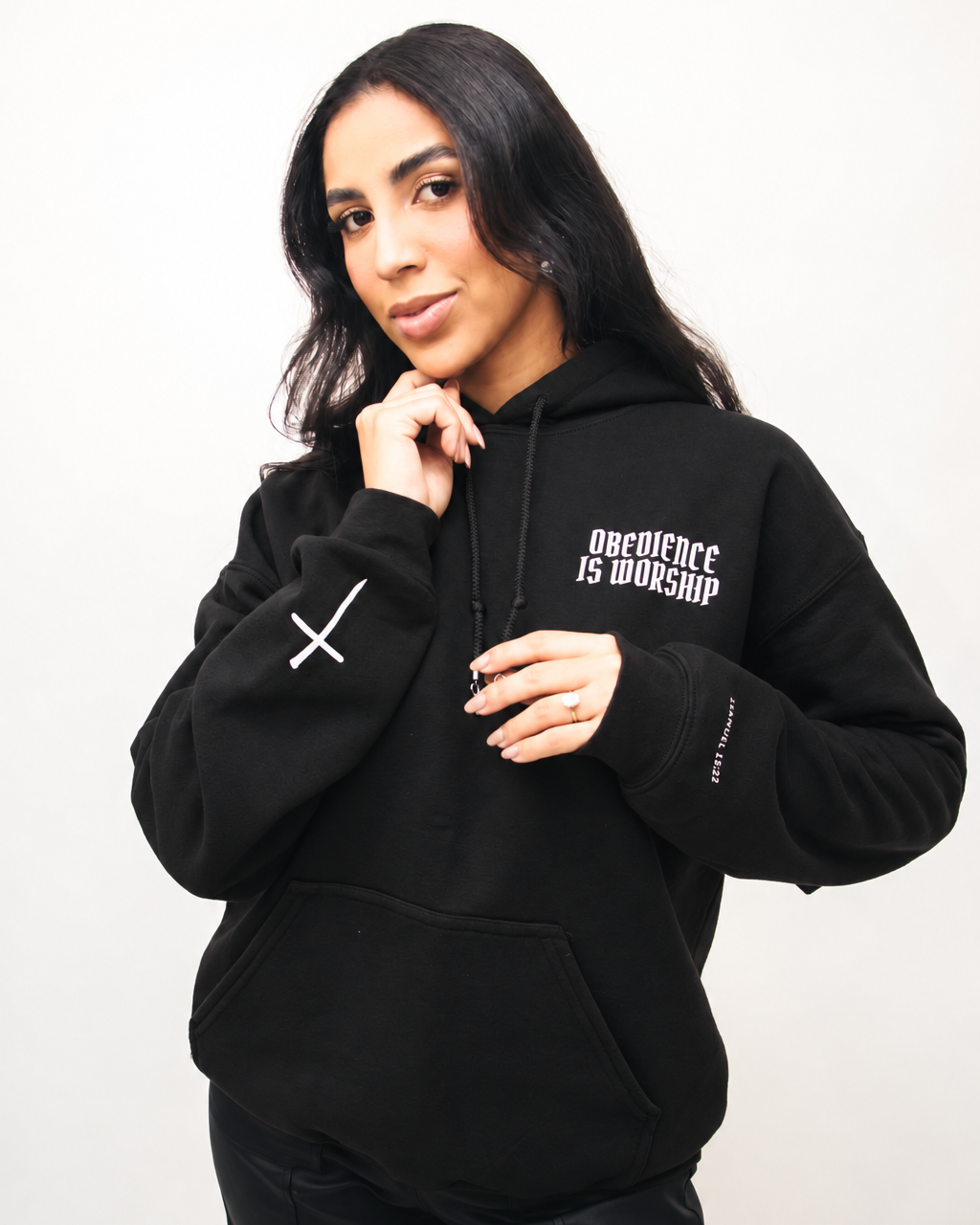 BLACK A woman wearing a black pullover hoodie with white text on the left chest reading 'Obedience is Worship' and a white cross graphic on the right sleeve, posed against a plain light background.
