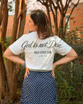 A woman photographed from behind outdoors, wearing a white graphic T-shirt with black script text reading 'God is not Done' and 'HOLY LOOKS CLUB', paired with a navy skirt with a small white geometric pattern.