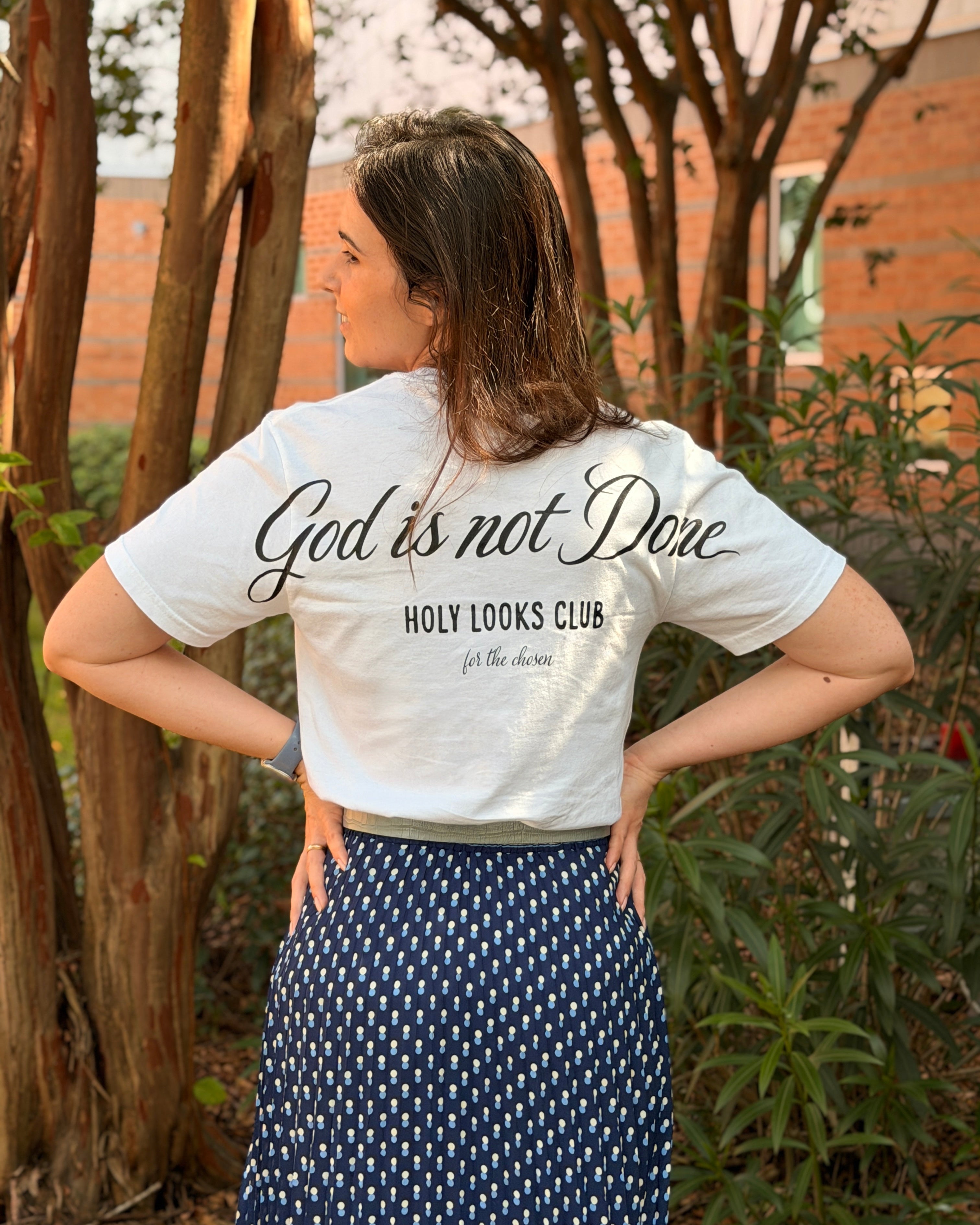 A woman photographed from behind outdoors, wearing a white graphic T-shirt with black script text reading 'God is not Done' and 'HOLY LOOKS CLUB', paired with a navy skirt with a small white geometric pattern.