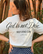 A woman photographed from behind outdoors, wearing a white graphic T-shirt with black script text reading 'God is not Done' and 'HOLY LOOKS CLUB', paired with a navy skirt with a small white geometric pattern.