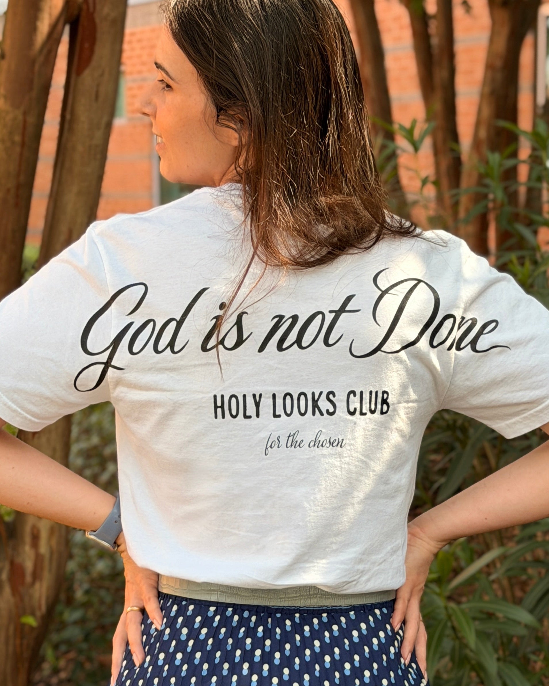 A woman photographed from behind outdoors, wearing a white graphic T-shirt with black script text reading 'God is not Done' and 'HOLY LOOKS CLUB', paired with a navy skirt with a small white geometric pattern.