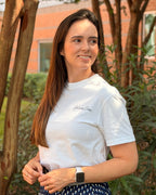 A woman standing outdoors in front of trees and a brick building, wearing a white short-sleeve t-shirt with a small script logo on the chest and a navy blue skirt with a small white geometric pattern.