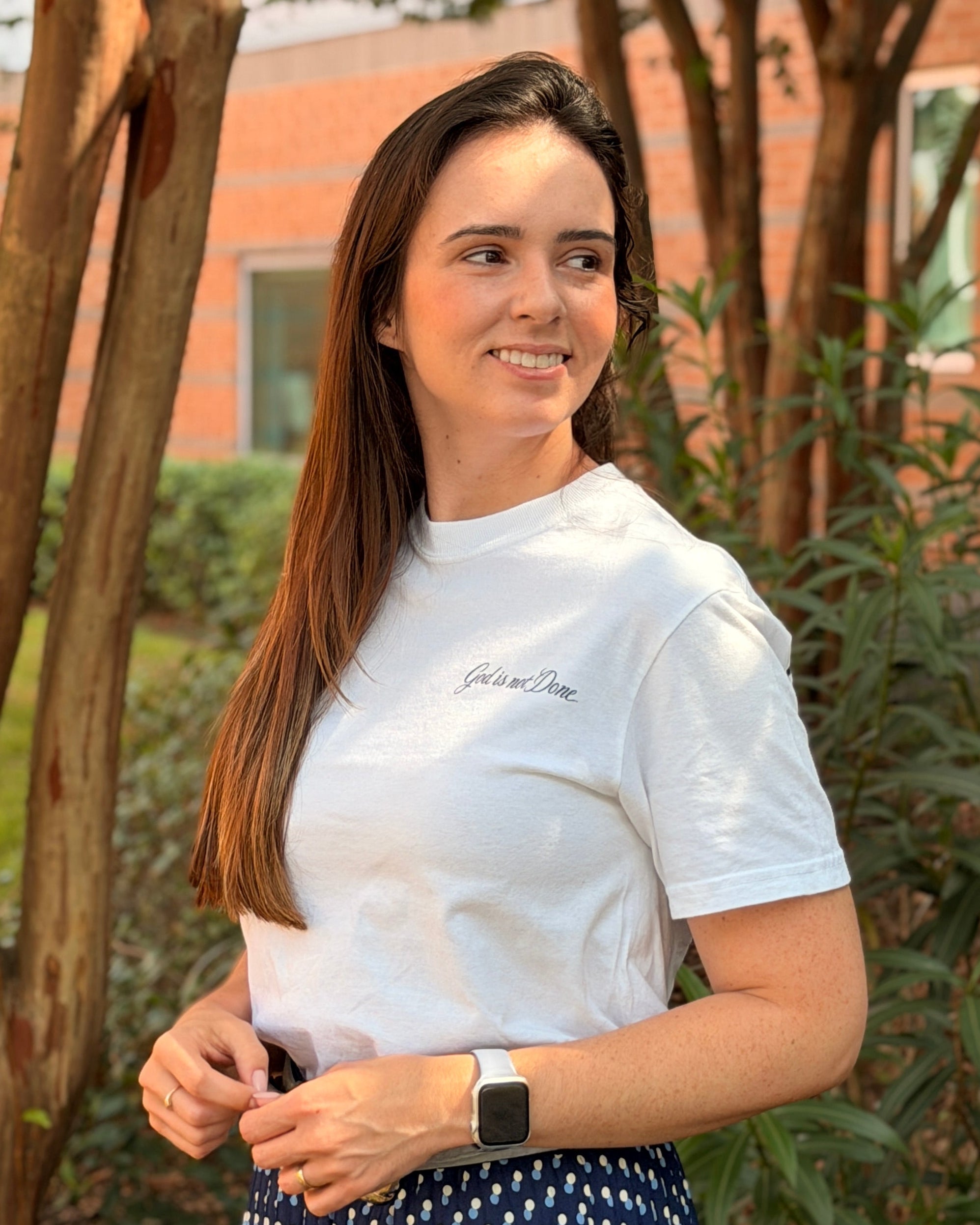 A woman standing outdoors in front of trees and a brick building, wearing a white short-sleeve t-shirt with a small script logo on the chest and a navy blue skirt with a small white geometric pattern.