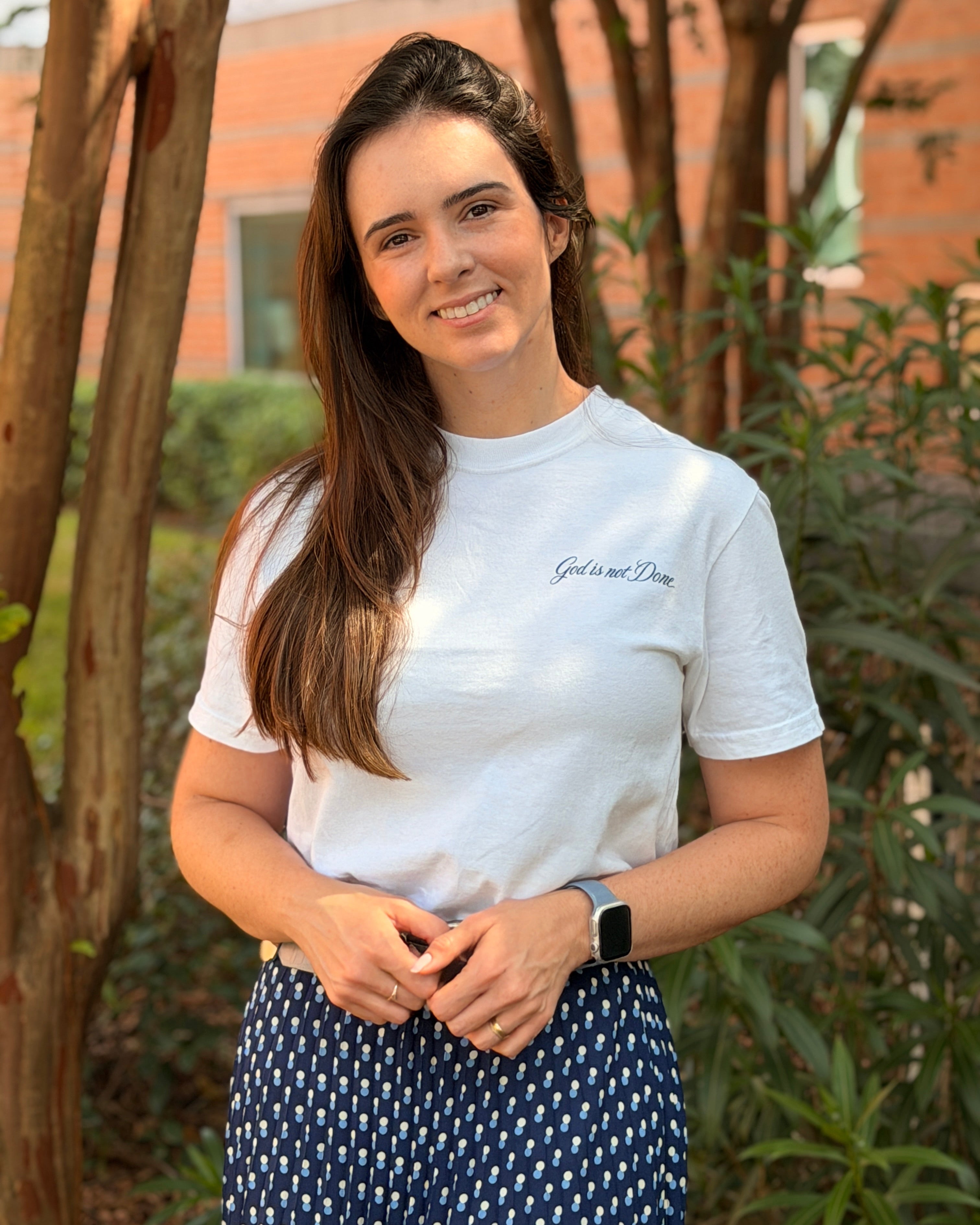 A woman with long brown hair stands outdoors wearing a white crew-neck t-shirt with 'God is not Disappointed' printed on the chest, paired with a navy blue and white patterned skirt.