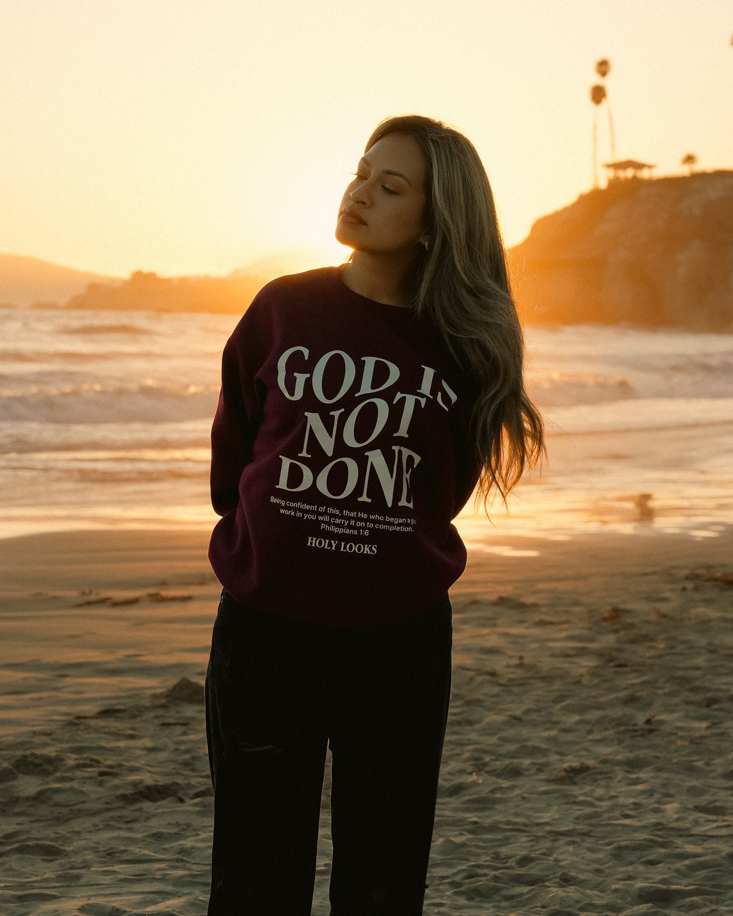 A woman stands on a beach at sunset wearing a dark maroon sweatshirt with the text 'GOD IS NOT DONE' in white, paired with black pants.