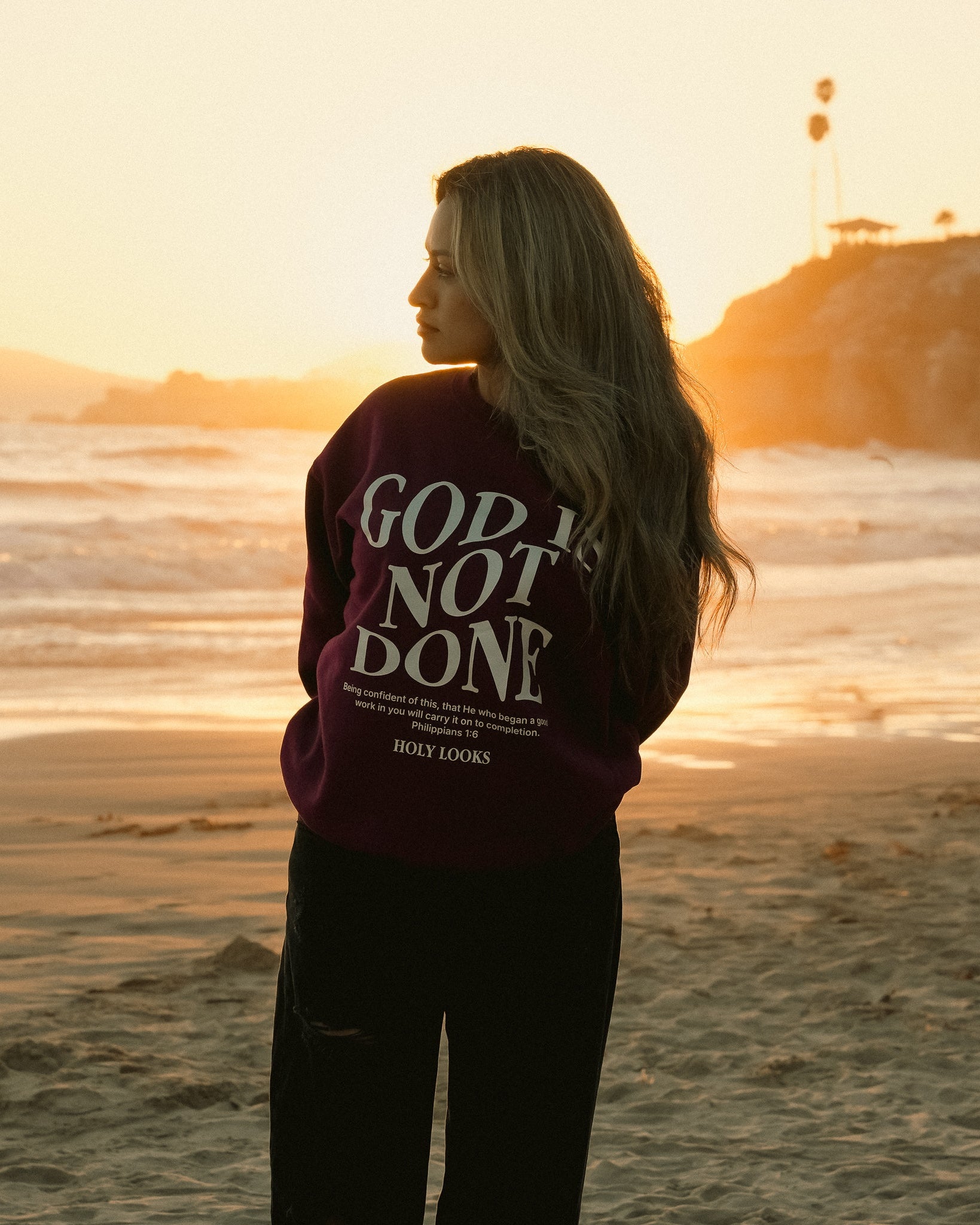 A woman stands on a beach at sunset wearing a dark plum sweatshirt with the phrase 'GOD IS NOT DONE' printed in large white letters on the back, paired with black pants.