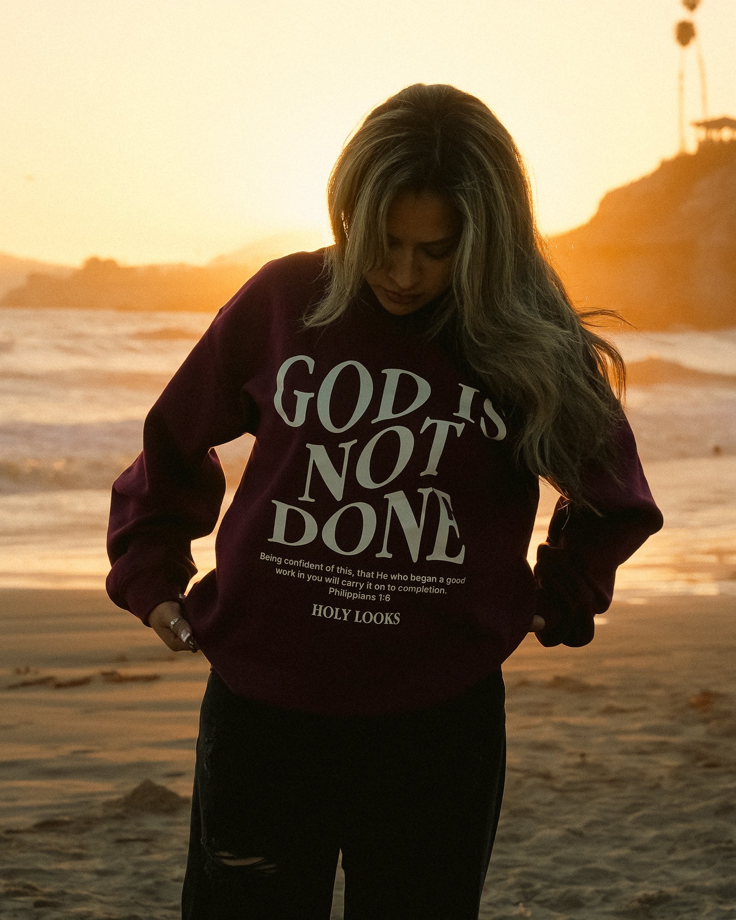 A woman stands on a beach at sunset wearing a dark purple sweatshirt with the text 'GOD IS NOT DONE' in white, paired with black pants.