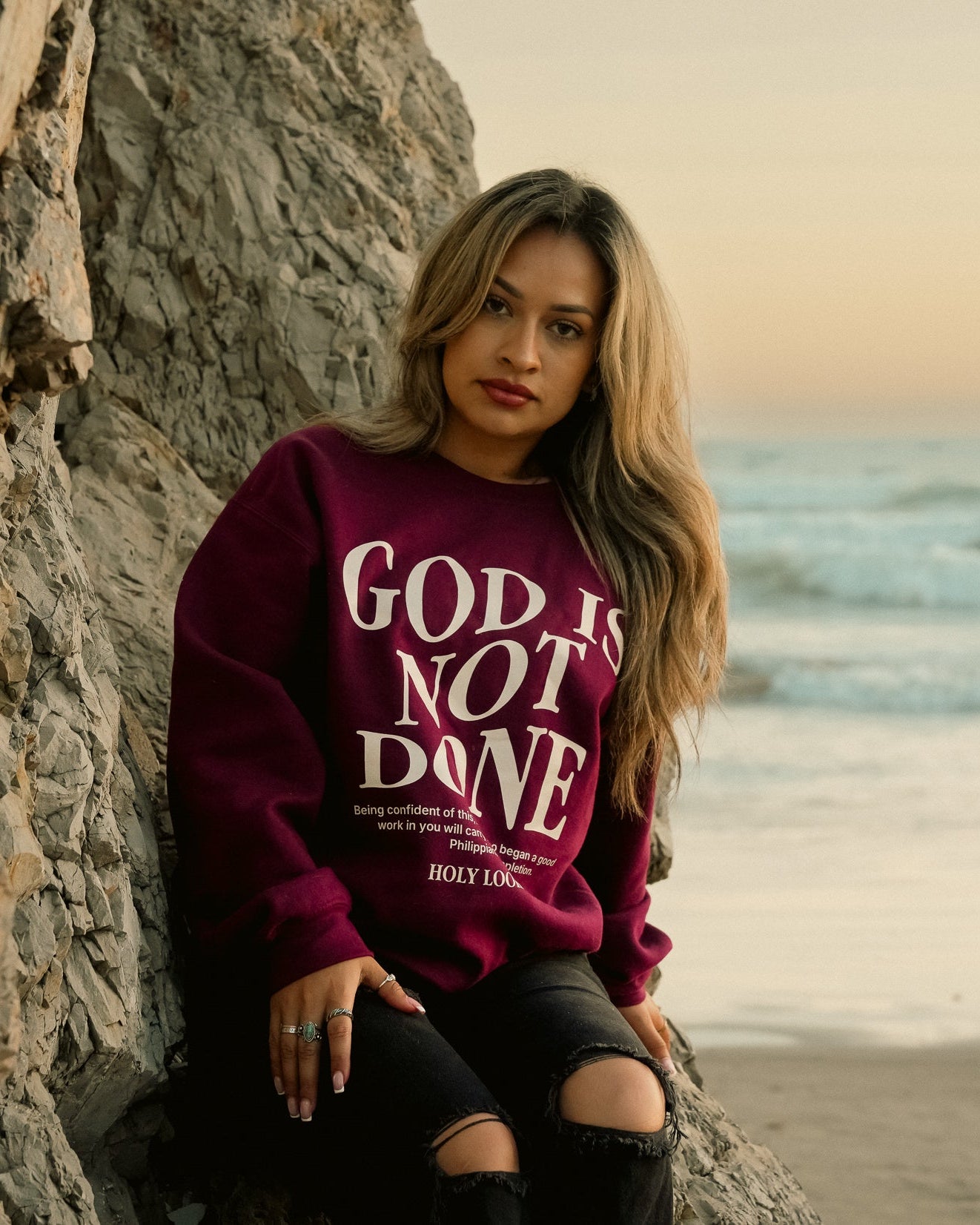 A woman sitting on a rock at the beach wearing a burgundy crewneck sweatshirt with the text 'GOD IS NOT DONE' in white, paired with black ripped jeans.