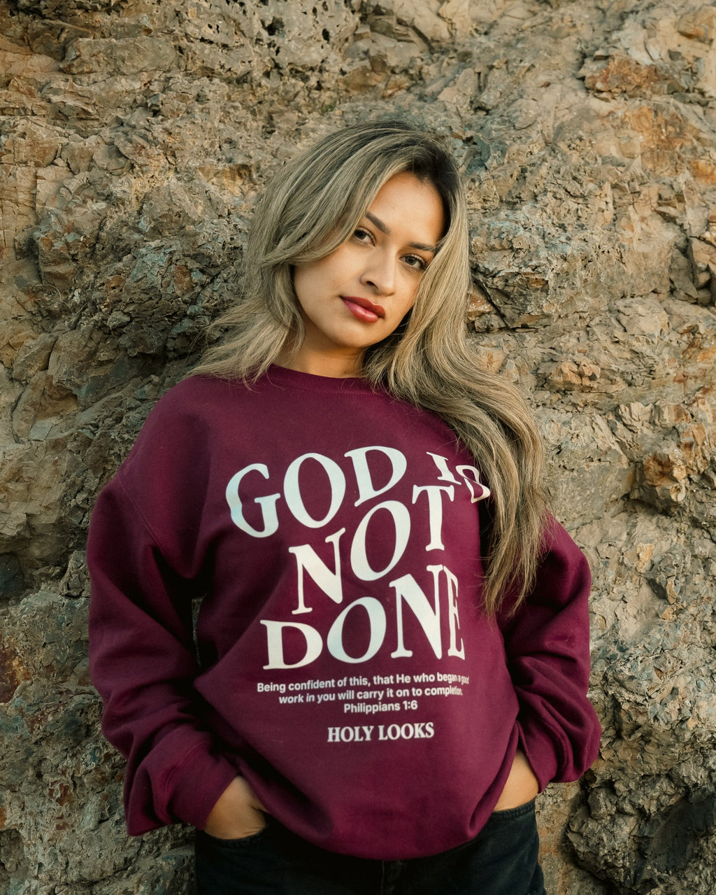A woman with blonde-highlighted hair stands against a textured stone wall wearing a maroon crewneck sweatshirt with the words 'GOD IS NOT DONE' in white text, paired with black pants.