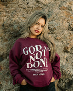 A woman with blonde-highlighted hair stands against a textured stone wall wearing a maroon crewneck sweatshirt with the words 'GOD IS NOT DONE' in white text, paired with black pants.